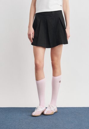 Femme portant une jupe plissée noire, un haut blanc, des chaussettes roses montant jusqu'aux genoux et des chaussures roses à lacets blancs, debout sur un tapis bleu.