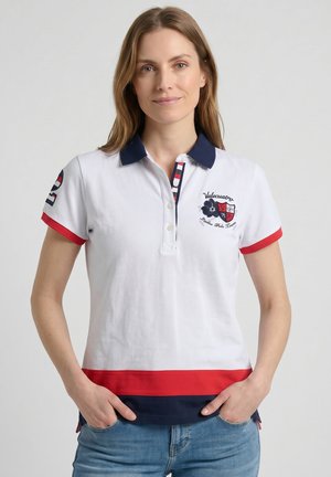 Mujer con polo blanco de cuello azul marino, ribetes rojos y azul marino, escudo bordado en el pecho y vaqueros azules, de pie con las manos en los bolsillos.