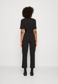 Combinaison noire à manches courtes, taille cintrée et pantalons à jambes larges. Présente un design épuré et est associée à des bottines noires.