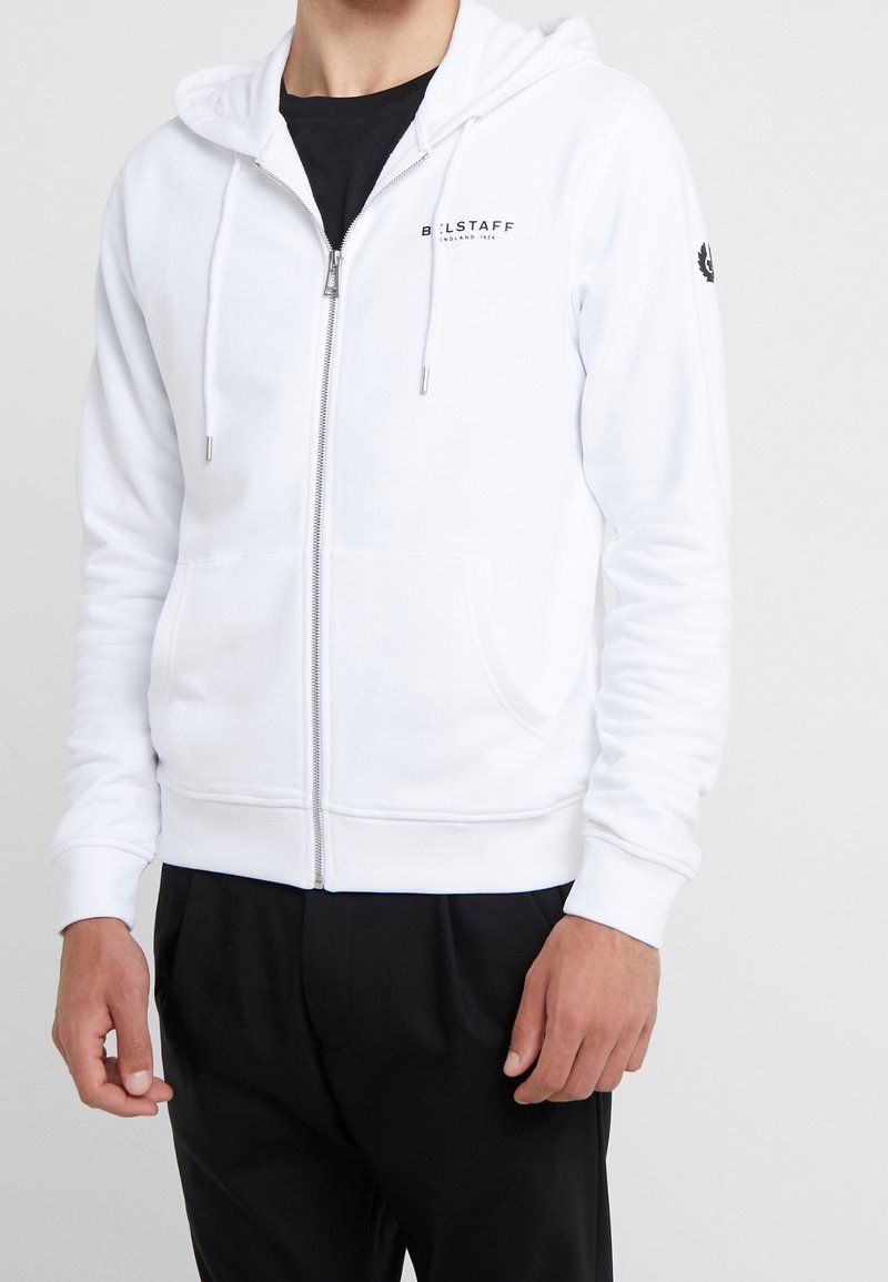 Homme portant un sweat à capuche blanc zippé avec une poche avant et un logo texte noir sur la poitrine, associé à un pantalon noir et un t-shirt noir en dessous.