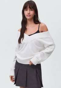 Bela široka pulover s dizajnom mimo ramen, teksturiranim V-izrezom, rebrastimi manšetami in robom, v kombinaciji s temno plisirano krilo.