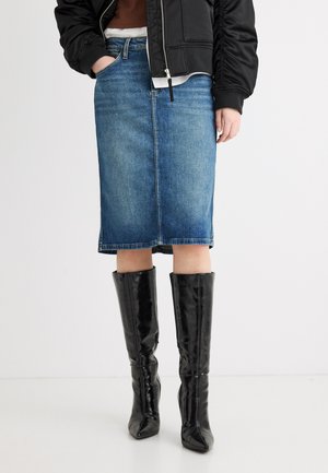 Ženska v modri denim krilu srednje dolžine, črnih škornjih do kolen in črni jakni, z roko v žepu.