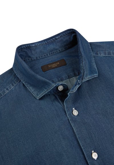 Camisa de mezclilla de manga larga con cuello abotonado y botones blancos, con una etiqueta marrón con "SLOWEAR" dentro del cuello.