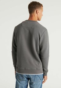 Uomo che indossa un maglione grigio a coste con maniche lunghe e jeans blu, in piedi con la schiena rivolta verso uno sfondo chiaro e neutro.