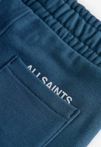 Tamno plave trenerke s teksturiranim materijalom, elastičnim pojasom i stražnjim džepom s ispisom "ALLSAINTS" u bijeloj boji.