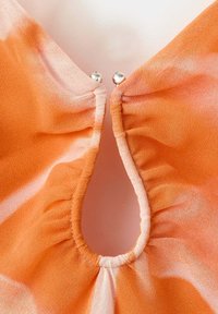 Gros plan sur un décolleté en tissu orange en forme de trou de serrure avec une petite fermeture à pression boule en argent en haut.