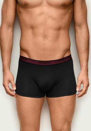 Männlicher Torso mit schwarzen Boxershorts und bordeauxfarbenem Bund, beschriftet mit "yamamay", vor neutralem Hintergrund.