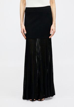 Γυναίκα που φοράει μακριά μαύρη φούστα με εφαρμοστή μίνι φούστα και διαφανές πλισέ ύφασμα που εκτείνεται μέχρι το πάτωμα.