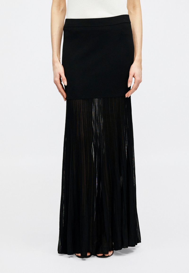 Γυναίκα που φοράει μακριά μαύρη φούστα με εφαρμοστή μίνι φούστα και διαφανές πλισέ ύφασμα που εκτείνεται μέχρι το πάτωμα.