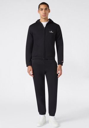 Man staat met een zwarte zip-up hoodie en bijpassende zwarte broek met witte sneakers op een witte achtergrond.