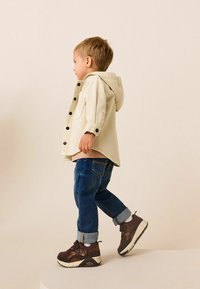 Lysbeige jakke med hætte, mørkeblå jeans og brune sneakers. Jakken har sorte knapper og opkrøllede ærmer. Enkel design.