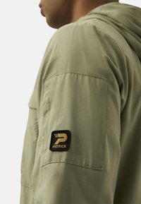 Green lightweight jacket with a hood, featuring a black and gold logo patch on the left sleeve and a pocket on the chest. Smooth texture.