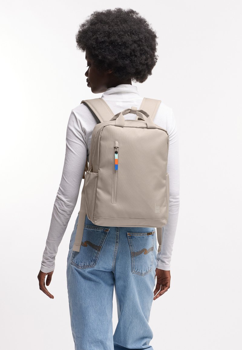 Persoon draagt licht beige rugzak met gekleurd ritsluiting, wit shirt met lange mouwen en blauwe spijkerbroek, van achteren tegen een witte achtergrond.