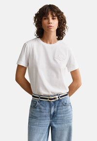 Femme aux cheveux bruns bouclés portant un t-shirt blanc avec un logo rentré dans un jean bleu avec une ceinture rayée noire et blanche, mains derrière le dos.