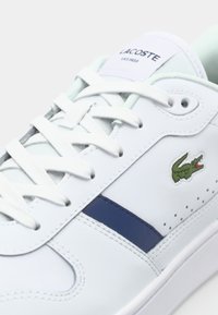 Zapatilla blanca de cuero con un logo de Lacoste en verde, una franja azul marino y cordones blancos. Textura suave con acentos perforados en los lados.