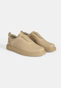 Beige instap schoenen met een geperforeerde textuur, een lichte hak en een gepolsterde binnenzijde. Heeft een gladde neus en-elastische zijkantpanelen.