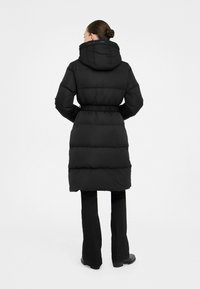 Cappotto imbottito nero con vita arricciata, cappuccio e colletto testurizzato, caratterizzato da un design trapuntato e maniche lunghe. Indossato sopra pantaloni neri.