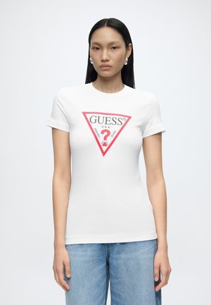Femme aux longs cheveux noirs portant des boucles d'oreilles créoles argentées, un t-shirt blanc Guess avec un logo triangle rouge et un jean bleu clair, debout devant un fond blanc.