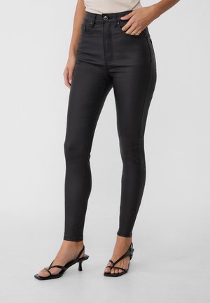 Femme portant un pantalon skinny noir taille haute et des sandales à talons petits noirs à lanières, debout devant un fond gris clair uni.