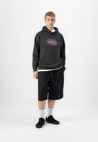 Jongeman staat in een zwart hoodie met een kleurrijk logo, zwarte knielange shorts, witte sokken en zwarte sneakers tegen een witte achtergrond.