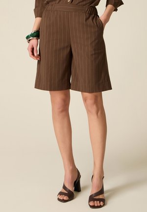 Brune knælange shorts med striber, båret med mørkebrune hælsandaler og et grønt armbånd på venstre håndled mod en beige baggrund.