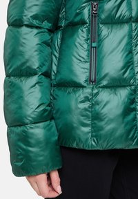 Zelená vycpaná bunda se rukávy a torzem, černým zipem, kterou nosí osoba v černých kalhotách, ruka uvolněná po straně.