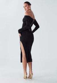 Melna kleita bez pleciem ar savilkumiem, garām pieguļošām piedurknēm un sānu šķēlumu. Aizmugurē ir aizdare ar rāvējslēdzēju. Kombinēta ar zelta augstpapēžu kurpēm.