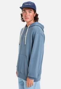 Sweat à capuche zippé en bleu clair, avec une poche avant, des cordons en crème et une coupe décontractée. Accompagné d'une casquette marine.