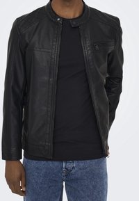 Zwarte leren jas met een drukknopkraag, doorgestikte schouderdetails en een voorvak met rits. Gedragen over een zwart shirt en blauwe jeans.