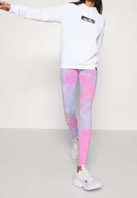 Bílá mikina s černým logem, spárovaná s legínami v růžovo-modrém tie-dye stylu a bílými sportovními teniskami. Hladký materiál, úzký střih.