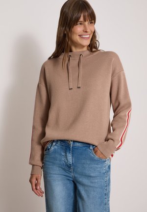 Kvinde smilende iført en lysebrun sweater med røde og hvide striber på ærmerne og blå jeans, stående med den ene hånd i lommen.
