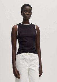 Schwarzes ärmelloses Top mit weißem Kragenbesatz, figurbetontes Design, kombiniert mit weißen Stoffhosen mit Kordelzug, die eine glatte Stoffoberfläche zeigen.