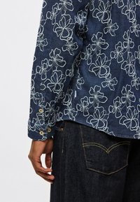 Πουκάμισο με μακριά μανίκια σε σκού ρο τζιν ύφασμα με λευκή κεντητή λουλουδένια διακόσμηση, διαθέτει μανίκια με κουμπιά και κυρτή ραφή στο κάτω μέρος, συνδυασμένο με παντελόνι από σκούρο τζιν ύφασμα.