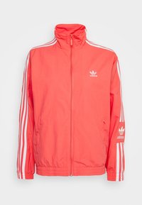 Korallrosa Adidas träningsjacka med vita tre-stripes detaljer på ärmarna, dragkedja fram, hög krage, och logotyp på bröstet och ärmen.