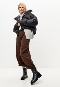 Mujer rubia vestida con una chaqueta de plumas negra, suéter gris, pantalones de pierna ancha marrones y botas negras hasta el tobillo, de pie contra un fondo blanco.
