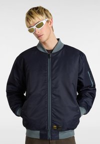 Marineblauer Bomberjacke mit Reißverschluss, hellgrauem Kragen und Bündchen, Seitentaschen und einem Etikett am unteren Saum. Getragen mit weißen Sonnenbrillen.