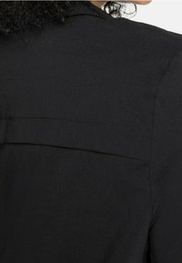 Person mit lockigem Haar, der eine schwarze Jacke trägt, die den oberen Rücken und den Schulterbereich zeigt, mit sichtbaren Nahtdetails.