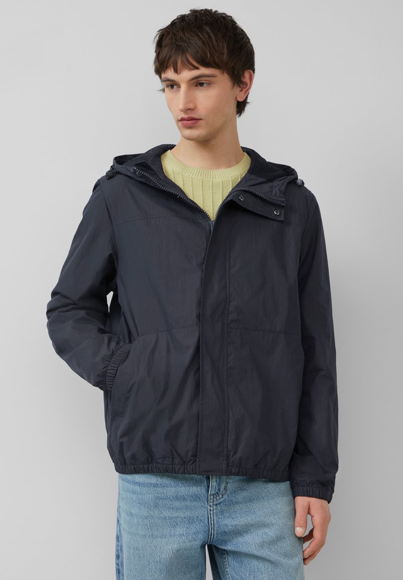 Marineblauwe lichtgewicht jas met een capuchon, elastische manchetten, twee voorzakken en een ritssluiting. Gemaakt van een gladde, waterafstotende stof.
