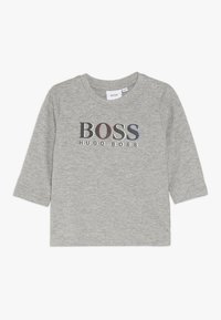 Graues Langarmshirt aus weichem Material mit einem auffälligen "BOSS"-Logo in Schwarz, Rot und Blau, mit Druckknöpfen an den Schultern für ein einfaches An- und Ausziehen.