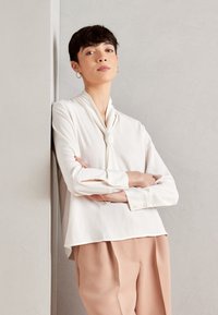 Bílá blůza s uzlovým detailem na krku, dlouhými rukávy s knoflíkovými manžetami, nošena s volnými kalhotami světle růžové barvy. Hladká textilie.