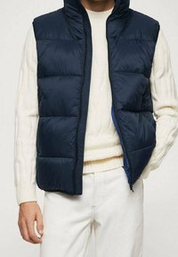 Laivaston sininen puhviliivi, jossa on korkea kaulus ja etuvetoketju. Varustettu vaakasuorilla tikatuilla paneeleilla ja sileällä, kiiltävällä pinnalla. Yksityiskohtana kermaharmaa neule.