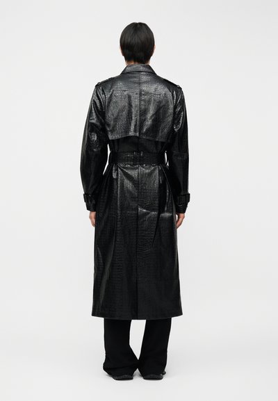 Άτομο που φαίνεται από πίσω, φοράει μακρύ μαύρο παλτό από συνθετικό δέρμα κροκοδείλου με ζώνη και μαύρο παντελόνι φαρδιάς γραμμής.