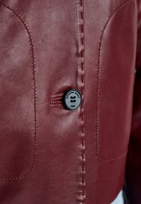 Nahaufnahme der Vorderseite einer kastanienbraunen Lederjacke mit schwarzem Knopf mit der Beschriftung „Milestone jackets 1994“ und detaillierter Naht.