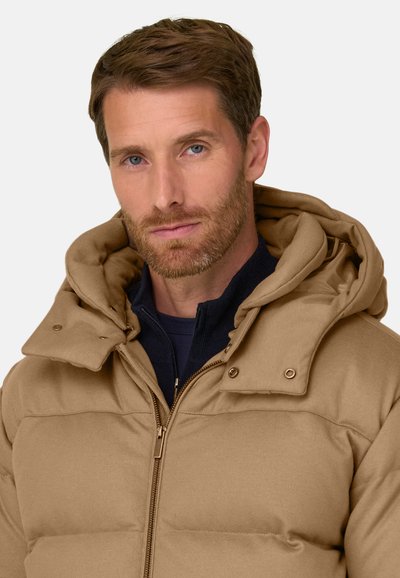 Homme aux cheveux bruns courts et barbe, portant une veste matelassée beige à capuche et un vêtement intérieur noir zippé sur fond blanc.