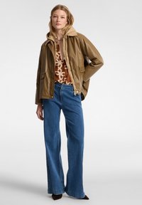 Giacca cappuccio marrone lucido, con due tasche frontali, indossata su un top marrone a fantasia. Abbinate a jeans azzurri a gamba larga e scarpe scure.