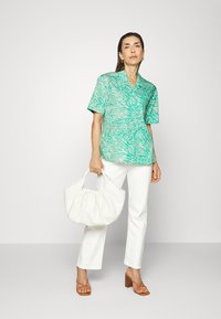 Camisa de manga curta verde e branca com riscas de zebra, combinada com calças brancas. Modelo segura uma mala branca e usa sandálias de salto em tom bege.