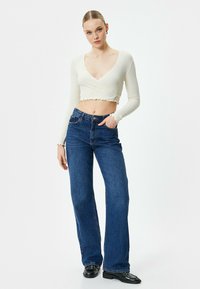 Hvid, ribbet langærmet crop top med v-hals og flæsekant, parret med højtaljede mørkeblå brede bukser. Sorte sko fuldender looket.