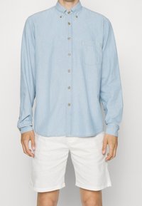 Jasnoniebieska koszula z długim rękawem, wykonana z denimu, z kieszonką na piersi i beżowymi guzikami. Połączona z białymi szortami.