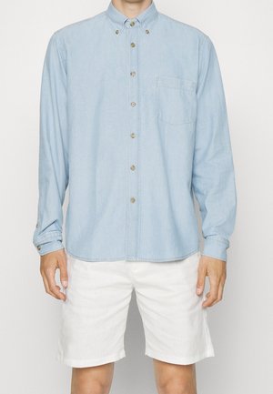 Camisa de mezclilla de color azul claro con botones, que presenta un bolsillo en el pecho, mangas largas y botones beige. Combinada con pantalones cortos blancos.