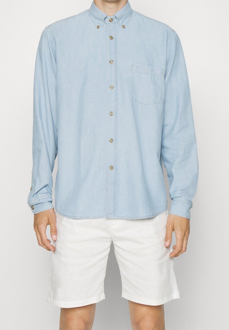 Jasnoniebieska koszula z długim rękawem, wykonana z denimu, z kieszonką na piersi i beżowymi guzikami. Połączona z białymi szortami.
